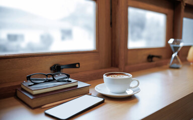 white coffee cup on wooden table by the window with mobile phone, book, eyeglasses and diary book for taking notes