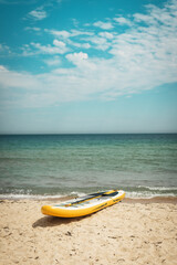 Bright yellow paddleboard lying on sandy beach near calm ocean. Clear sky and peaceful waves evoke summer freedom, sport activity, seaside vacation atmosphere. Perfect for travel and lifestyle themes.
