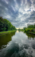 A tranquil river scene peacefully reflecting the vibrant lush greenery and striking dynamic cloud...