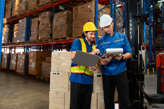 Warehouse supervisor and worker discussing inventory checklist using laptop and clipboard in large distribution center. Efficient teamwork in industrial supply chain logistics.