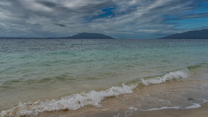 Tropical seascape. Turquoise ocean waves are foaming on a secluded sandy beach. Mountains on the horizon. Blue sky, clouds. Madagascar.