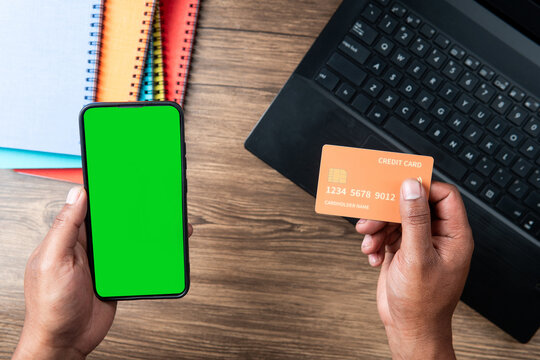 Hand holding a credit card and smartphone with green screen in front of laptop on wooden desk, perfect for illustrating online payment, e-commerce, digital transactions, and fintech concepts. - Powered by Adobe