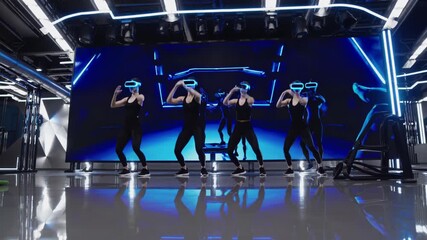 Female athletes wearing vr headsets performing synchronized fitness exercises in futuristic gym with digital virtual instructor on large screen