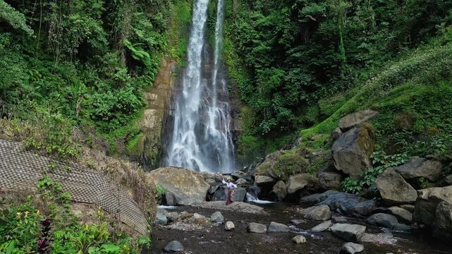 A Balinese female dancer performs gracefully in nature, wearing traditional attire and a leaf headpiece. Surrounded by greenery and shrubs, with Gitgit Waterfall visible in the background.
