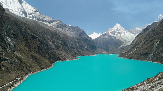 A spectacular aerial shot pans alongside a steep mountain, revealing the vast, vibrant turquoise waters of Laguna Par&oacute;n and the snow-capped peaks of the Cordillera Blanca in Peru.