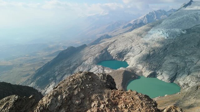 A dramatic aerial shot pulls back from two stunning turquoise alpine lakes and a vast glacier to reveal a rocky mountain ridge at Paso del Zorro in the remote Huayhuash range of Peru.