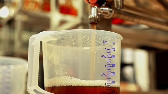 A close-up shows fermented kombucha tea being poured from a tap into a large measuring pitcher during the commercial brewing and bottling process, highlighting the healthy probiotic drink.
