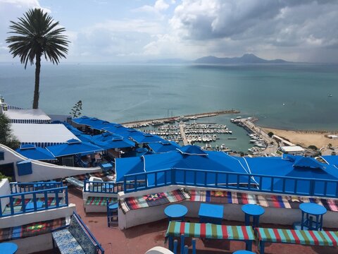 A scenic view from Sidi Bou Said, overlooking the marina and Mediterranean Sea. Blue and white terraces with colorful cushions contrast beautifully with the turquoise water and distant mountains