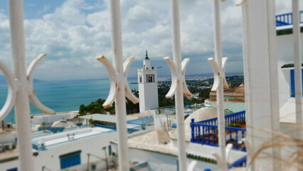A scenic view of a white coastal town through decorative iron bars, featuring a tall minaret, blue balconies, and the turquoise sea stretching to the horizon under a partly cloudy sky.