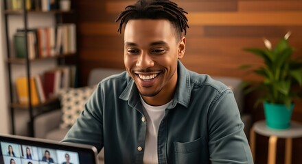 Fototapeta premium Young man working on a laptop, smiling happily while participating in an online meeting at home with a cozy atmosphere.