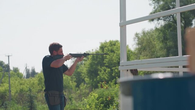 close up drum in foreground, side view of man aiming rifle toward distant target near farm frame, rural greenery background, protective action to guard crops from pests