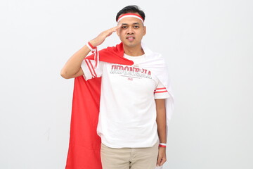 Naklejka premium Indonesian man with red and white ribbon to celebrate Indonesian independence day standing with hand gesture saluting, standing isolated on white background.