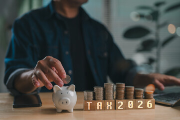 Person plans future 2026 tax payments saving money. Their hand puts coin piggy bank beside growing coin stacks, symbolizing financial planning, investment, future budgeting.