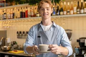 Smiling male barista in apron holding cup standing at counter in modern coffee shop interior
