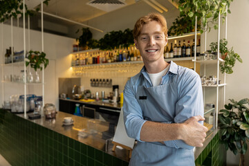 Cheerful young café worker with crossed arms leaning against counter in stylish interior