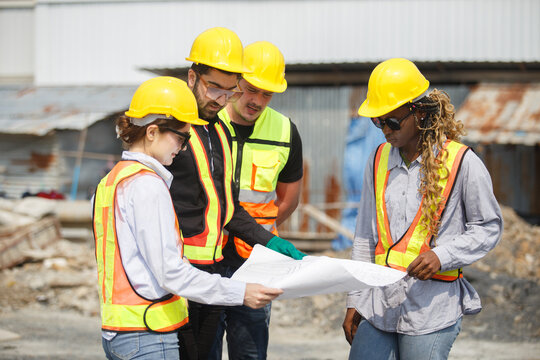 Diverse team of construction workers wearing safety helmets and reflective vests looking blueprint discussing building plans on construction site for teamwork and project planning concepts.