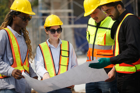 Diverse team of construction workers wearing safety helmets and reflective vests looking blueprint discussing building plans on construction site for teamwork and project planning concepts.