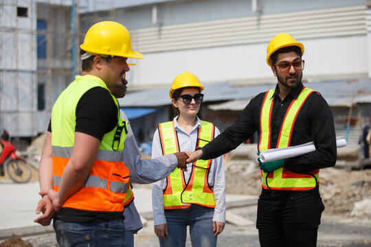 Diverse construction team wearing safety helmets and reflective vests shaking hands at building site symbolizing teamwork, agreement, partnership and successful project collaboration.
