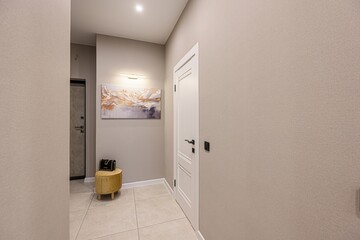 modern hallway with beige walls, tiled floors, a white door, artwork, and a yellow stool with a black bag