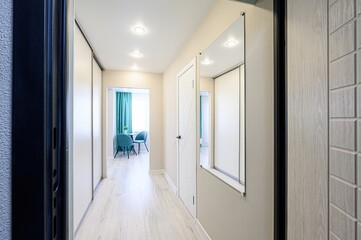 bright hallway with wooden flooring, sliding door wardrobe, a white door, and a glimpse into a room with blue chairs and curtains. A mirror reflects the space