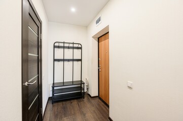 bright hallway with wooden floors. Features include a dark wood door, a metal coat rack with a bench, and a light-colored front door, all against white walls