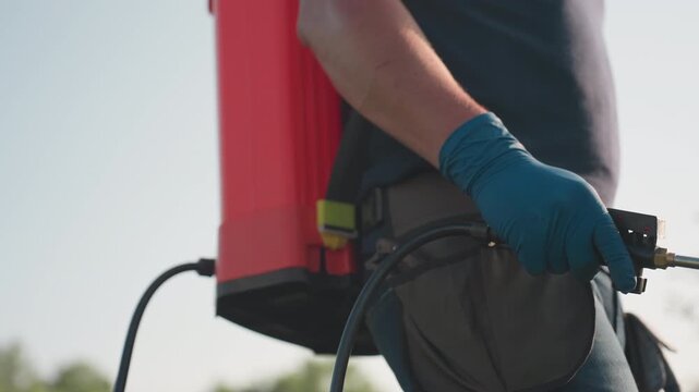 close up of man fumigating field with backpack sprayer wearing glove and protective gear, sunlight highlighting arm and hose during outdoor agriculture pest control