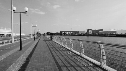 Naklejka premium A black-and-white riverside promenade with modern lamp posts, railings casting shadows on the walkway, and industrial buildings in the distance under a slightly cloudy sky. 