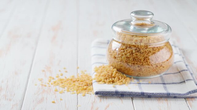 Dry bulgur grains in glass storage jar on a white wooden table.
Raw bulgur in a storage glass jar on a light kitchen table. 