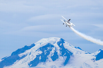 Wing walker on a vintage biplane climbs above a snow-capped mountain, white smoke streaming across a blue sky, a dramatic aerobatics symbolizing courage, freedom and high altitude adventure.