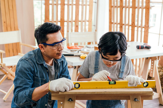 Father mentors son in carpentry, teaching precise measurements with a spirit level. Both wear safety glasses and gloves, highlighting teamwork, skill, and family craftsmanship in a bright workshop.