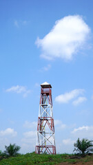 Metal observation tower rising against a vibrant blue sky, encircled by lush greenery and palm trees, highlighting rural industrial infrastructure