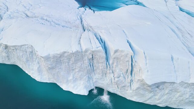 A tall melting iceberg in the Arctic Ocean. A glacier with a lake and waterfall. Clear ice floats in turquoise water. Ecology, melting ice, climate change, global warming. Aerial view. Midnight sun.