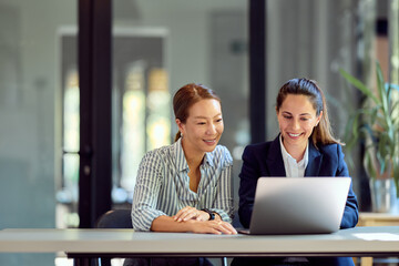 Two Businesswomen Collaborating and Smiling While Using a Laptop at an Office Desk