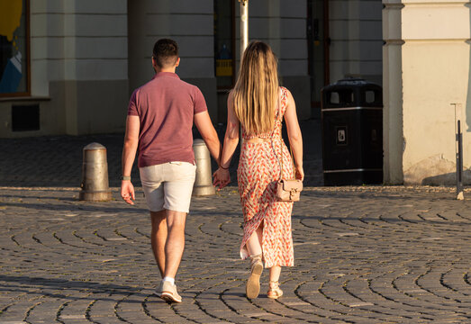Rear view of young couple walks hand in hand on a cobblestone street at sunset, dressed in summer clothes and bathed in warm golden light. Stylish couple in love walking on the street, holding hands  - Powered by Adobe