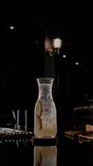 A tall glass bottle of iced lemon water with chia seeds, placed on a round wooden coaster against a dark background with artistic smoke swirling around, creating a dramatic and refreshing presentation