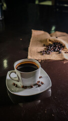 A white ceramic cup filled with rich black coffee, placed on a square white saucer with scattered coffee beans, set on a dark reflective surface with a blurred background of coffee beans on burlap.