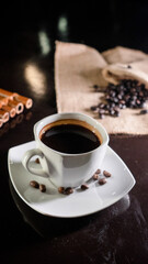 A white ceramic cup filled with rich black coffee, placed on a square white saucer with scattered coffee beans, set on a dark reflective surface with a blurred background of coffee beans on burlap.