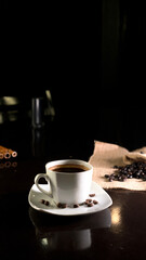 A white ceramic cup filled with rich black coffee, placed on a square white saucer with scattered coffee beans, set on a dark reflective surface with a blurred background of coffee beans on burlap.