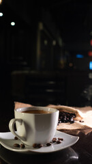 A white ceramic cup filled with rich black coffee, placed on a square white saucer with scattered coffee beans, set on a dark reflective surface with a blurred background of coffee beans on burlap.