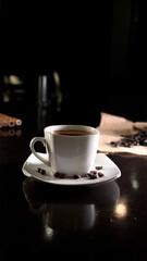 A white ceramic cup filled with rich black coffee, placed on a square white saucer with scattered coffee beans, set on a dark reflective surface with a blurred background of coffee beans on burlap.