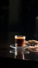 A clear glass cup filled with rich black coffee, placed on a transparent saucer over a dark reflective surface, with scattered coffee beans and a burlap cloth in the blurred background.
