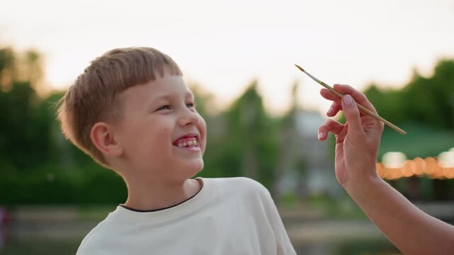 close up teenage boy turning face away as playful mom teases with paint brush by pool under sunset glow, boy smiles while dodging gentle stroke capturing warm evening bonding moment on wooden dock