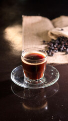 A clear glass cup filled with rich black coffee, placed on a transparent saucer over a dark reflective surface, with scattered coffee beans and a burlap cloth in the blurred background.