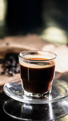 A clear glass cup filled with rich black coffee, placed on a transparent saucer over a dark reflective surface, with scattered coffee beans and a burlap cloth in the blurred background.