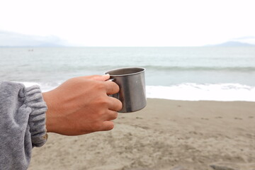 A person's hand holding a metal travel mug on a tranquil beach by the sea