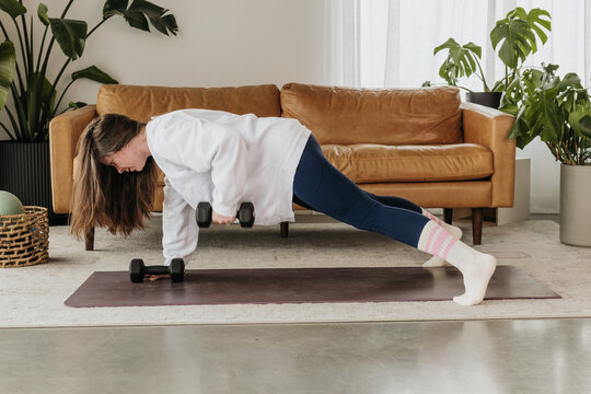 Woman lifts weights while in plank on yoga mat in living room