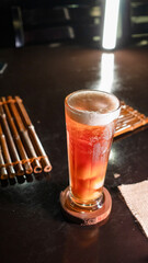 A tall glass of iced lychee tea with a vibrant orange gradient, containing whole lychee fruits at the base, served on a wooden coaster over a dark reflective surface, creating a refreshing and tropica