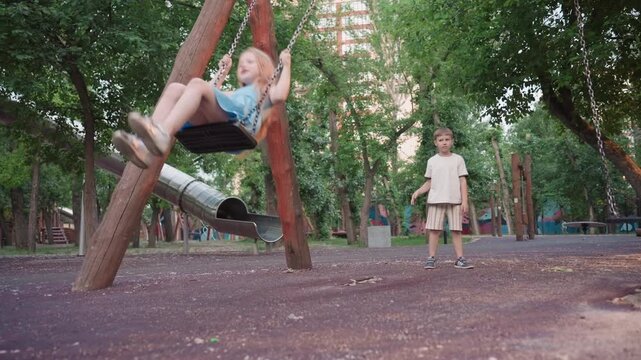 teenage girl smiles as she swings on wooden chain swing while playmate pushes from behind in lush park at dusk capturing joyful sibling play under warm evening sunlight shimmer