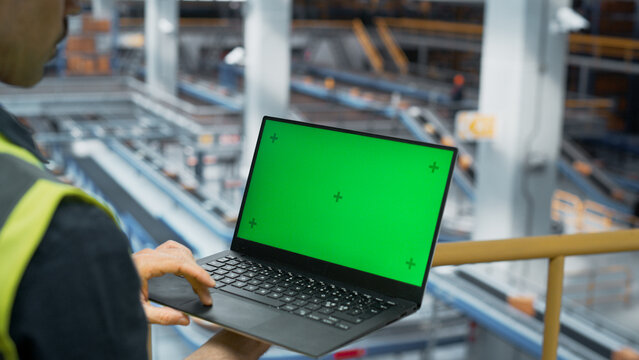 Male Employee Working at Modern Logistics Center With Conveyor Belt. International Online Shopping Business Facility. Caucasian Man Using Laptop Computer with a Green Screen Chromakey Mock Up Display. - Powered by Adobe