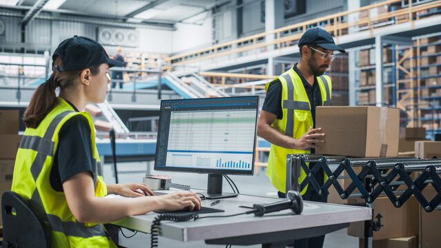 Caucasian Female Stocking Associate Using Desktop Computer To Check Inventory In Modern Distribution Facility. Multiethnic Colleague Loading Cardboard Boxes With Orders On Automated Conveyor Belt